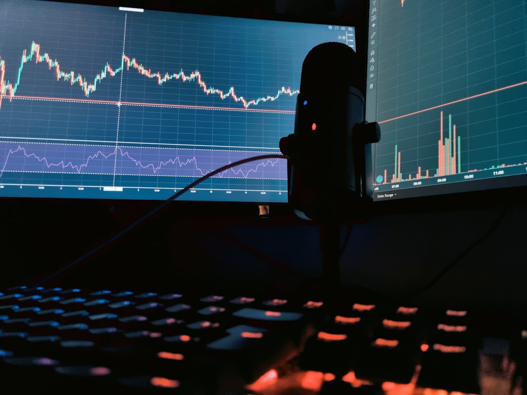 A display of two cryptocurrency markets on different monitors with a microphone and multicoloured keyboard in the foreground.